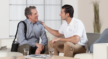 Homecare, Oxygen patient with nasal cannula sitting on a couch in a living room at home carrying the device around his shoulder,  sitting together with male nurse who touches his shoulder